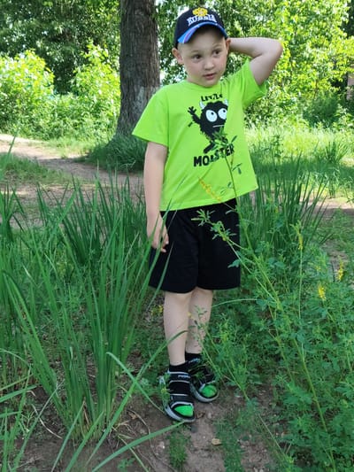 In the city of Raikov, Russia, a young boy stands amidst lush greenery. He is wearing a bright neon green t-shirt with a cartoon monster graphic and the word "MONSTER" printed below it, paired with black shorts. A dark baseball cap with "RUSSIA" written on it adorns his head. His right arm is raised, touching the back of his head. He wears black socks with "POWER" written on them and black and green sneakers. The environment is a natural setting, likely a park or wooded area, with tall grass and trees in the background. The ground appears to be a dirt path.