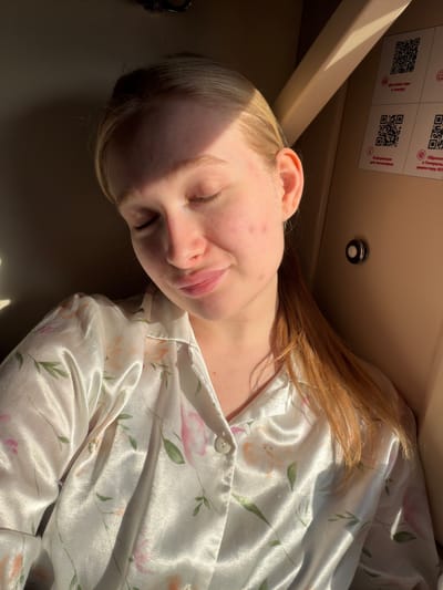 A fair-skinned young person lies in an enclosed space, possibly a compartment. The person has blonde hair with reddish-brown streaks, closed eyes, and slightly parted lips. The person's skin has some redness and small blemishes. They are wearing a light-colored satin-like garment with a floral pattern. The background includes parts of a ceiling with a small light fixture and labels with QR codes. The image was taken in a compartment in Krasnyy Sulin, Russia.