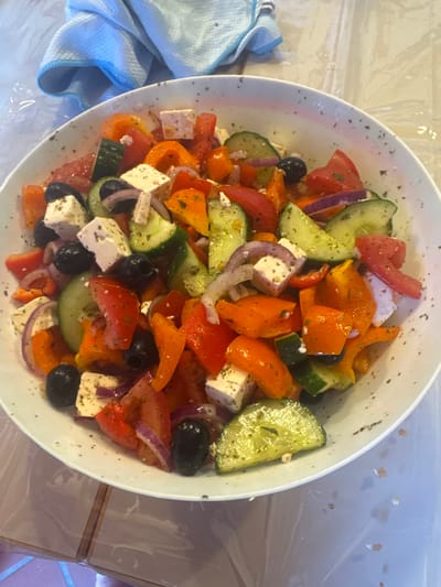 An overhead, close-up shot shows a white bowl filled with a Greek salad. The salad contains cubed feta cheese, chopped tomatoes, sliced cucumbers, black olives, red onion wedges, and orange bell pepper pieces. The ingredients are mixed and sprinkled with dried herbs. The bowl is placed on a light-colored, reflective surface. In the upper left corner, a crumpled light blue towel is visible. The background is out of focus and appears to be a wall with tiles. The scene does not provide any information about the city of Merzenich, Germany.