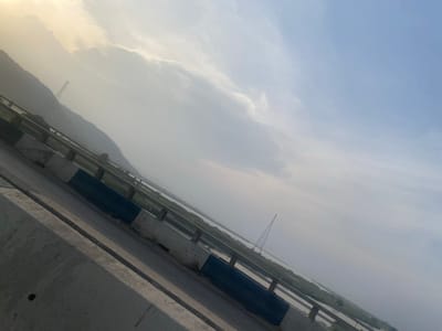 The image presents a panoramic landscape captured from an elevated perspective, likely a bridge or overpass. The scene, when viewed upright, shows a wide expanse of sky and distant land, with elements of the bridge structure visible in the foreground.
In the upper portion of the image, the concrete structure of a bridge or elevated roadway is visible, featuring a textured surface and horizontal lines. A metal railing or barrier runs along the edge of this structure, providing a clear demarcation. Some dark blue or black rectangular panels or markings are affixed to or painted on the concrete elements of the bridge.
Below the bridge, a vast body of water, possibly a river or large lake, stretches across the midground. On the far bank of this water, there appears to be flat, low-lying terrain, which then transitions into a range of distant mountains or hills that rise softly against the horizon. The atmosphere over the water and land is hazy, creating a sense of depth and distance.
A prominent feature in the mid-left distance is a tall, triangular lattice structure, characteristic of a power transmission tower. Another, smaller such structure might be faintly discernible near the base of the distant mountains on the right.
The sky occupies a significant portion of the image, displaying a gradient of colors from a light blue at the top to a softer, warmer, and more yellowish hue near the horizon, especially towards the right side. Scattered, soft white clouds are visible across the sky, suggesting a partly cloudy day. The soft, diffused light and the warm glow near the horizon indicate either early morning or late afternoon/evening.
The overall impression is one of tranquility and vastness, with a blend of natural landscape and man-made structures.