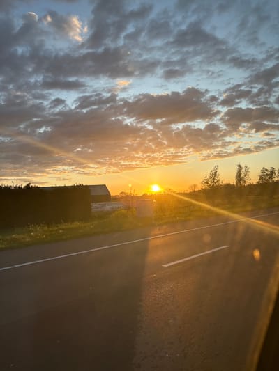 The image captures a sunset scene taken from a moving vehicle, possibly on a highway. The road stretches to the right of the frame with a white lane marking. Adjacent to the road, there's a grassy verge with a dark, bushy area.

The sunset is prominent, casting a golden glow on the clouds and the landscape. The sky is a mix of blue and orange hues, with sunlight streaming through the clouds.

The location cannot be identified or inferred.