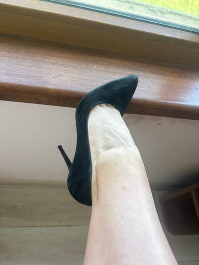 A close-up, vertical shot shows a person's right foot wearing a black high-heeled shoe. The shoe is a pointed-toe pump with a stiletto heel, made of a textured material that appears to be embossed leather. The shoe is positioned on what looks like a wooden surface, possibly a windowsill or shelf, adjacent to a window with a bright green, out-of-focus view outside. The foot is resting inside the shoe, with the arch and the top of the foot visible. The skin on the foot and lower leg shows some texture and visible veins. The background consists of a light-colored wall and further wooden paneling. The lighting suggests it is daytime, likely indoors. The image is taken in Prodanovtsi, Bulgaria. There is no visible text in the image.