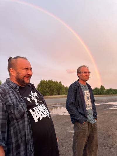 This is a photograph of two men standing outdoors with a rainbow in the sky. The man in the foreground, on the right side of the image, has a beard and is wearing a black t-shirt with white text that reads "LINKIN PARK" and a grey and black plaid shirt. He appears to be squinting or wincing. The man in the background, on the left, is wearing glasses, a blue patterned jacket, and a light blue t-shirt. He has a cigarette in his mouth and is looking upwards. The setting appears to be a paved area, possibly a parking lot, with trees and a road visible in the background. The sky is overcast, with a visible rainbow indicating that it may have rained or is about to rain. The lighting suggests it is either early morning or late afternoon. The overall mood seems casual and candid, with the subjects reacting to the natural phenomenon of the rainbow. The location is identified as Sovkhoznyi, Russia.