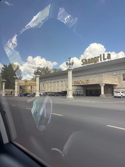 The image depicts a view from inside a car, oriented towards the Shangri La Casino in Verin Ptghni, Armenia. The camera is positioned from the front passenger seat. The casino building is visible in the middle of the frame. Cars are parked in front of the casino. The sky is blue with white clouds. The reflection on the windshield is distracting.