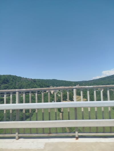 The image shows a view from a bridge over a green, hilly landscape. The sky is clear and blue. The bridge has a metal railing in the foreground. In the middle ground, there are trees and some buildings, suggesting a village. The scene is likely the village of Villards-d'Héria, France, which is known for its picturesque scenery and bridges. The image is taken from a moving vehicle, as evidenced by the slight blurriness.