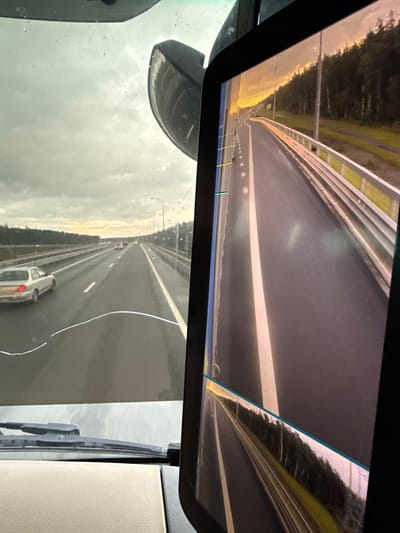 The image shows a view from inside a vehicle, looking out through the windshield and at a display screen. The windshield shows a highway with several cars and a cloudy sky. The display screen shows a view of the highway from the perspective of a rear-view camera, with a guardrail and a forest on the side of the road. The sky is illuminated by the setting sun, casting a warm glow on the scene. The location is identified as the city of Priladozhsky, Russia. The scene appears to be a typical highway drive, possibly during evening rush hour or a journey through a rural area. There are no people clearly visible in the image, and no specific actions or interactions are depicted. The environment is characterized by a highway, vehicles, trees, and a cloudy sky.