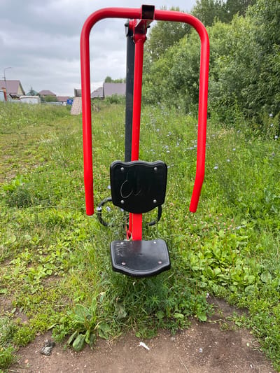 This is a full, eye-level, outdoor shot of a red and black outdoor fitness machine situated in a grassy field. The machine has a black seat and backrest. The red frame consists of two curved upright bars extending from a central black pole. The background features a mix of green trees and vegetation, with a few houses and a cloudy sky visible in the distance. The location is identified as Nagayevo, Russia. The ground around the machine is dirt and grass, with some wildflowers scattered throughout.