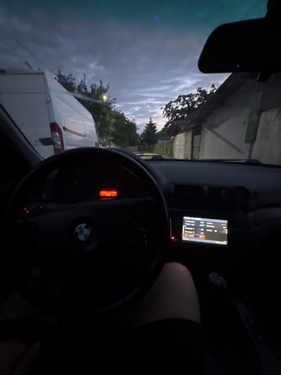 The image depicts the interior of a vehicle, likely a BMW based on the steering wheel emblem. The view is from the driver's perspective, showing the steering wheel, dashboard with a digital display showing "235551 - 3985 300", and a central infotainment screen. The infotainment screen displays vehicle information including "Autonomia 300 Arrivo", "Temp. ext. 28.0° Distanza 412", "Consumo 1 4.5", "Consumo 2 4.8", and "Vel. medio 28.0". The date "08.04.2020" is also visible on the screen. Outside the vehicle, a white van is parked to the left, and a building with a fence is visible to the right, suggesting an urban or semi-urban environment. The sky appears overcast, indicating it is either early morning or late evening. This scene is situated in the city of Straldzha, Bulgaria.