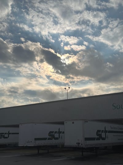 A bright, iridescent cloud formation with the sun peeking through is visible in a grey and blue sky above a long, low-rise industrial building. The building has a light-colored facade, and a sign with the word "Sou" is partially visible on the right side. In front of the building, several white semi-trailer trucks are parked. The trucks have green lettering that appears to be a logo and the words "SOUTHERN CHAMPION". The scene is likely an industrial area or warehouse in Chattanooga, United States of America.