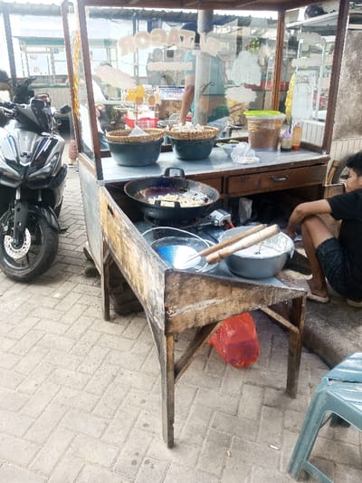 A street food vendor's cart features a fryer with cooking items, baskets of fried goods, and condiments. A person sits nearby. A motorcycle is parked next to the cart. The cart's construction appears basic, with exposed wood and a glass display case. A red plastic bag hangs below. A plastic chair is visible. The background includes a busy street scene. The scene suggests an outdoor dining or takeaway food establishment.