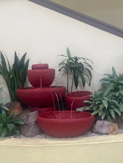 The image shows a three-tiered red fountain, likely ceramic or terracotta, with water flowing from one level to the next. The fountain is surrounded by various green plants and stones, creating a decorative and naturalistic arrangement. The entire setup appears to be placed against a wall, with the top left corner of the image showing a portion of the wall and trim. The lighting suggests an indoor environment. There is no visible information that confirms this is Lilongwe, Malawi.