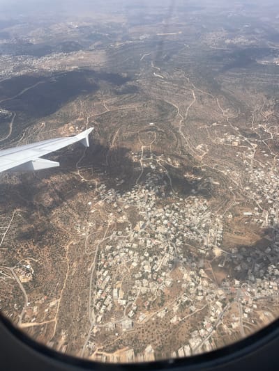 An aerial view captures the landscape surrounding the Palestinian town of Kobar, located in the West Bank. The image showcases a terrain characterized by rolling hills, sparsely vegetated with shrubs and scattered trees, likely olive groves. Numerous winding dirt roads and paved pathways crisscross the landscape, connecting various clusters of buildings. These settlements consist primarily of small to medium-sized houses with light-colored roofs, indicative of rural or semi-rural residential areas.
In the foreground, the wing of an airplane is visible, extending from the lower left corner and cutting across the upper left quadrant of the image. The wing is white and appears to be in good condition. The airplane window framing is also visible along the bottom edge of the photograph.
The overall impression is one of a developing landscape, with human habitation interspersed within a natural, arid environment. The presence of the airplane suggests the photograph was taken during a flight over the region. The light suggests it is daytime, with some atmospheric haze visible in the distance.
