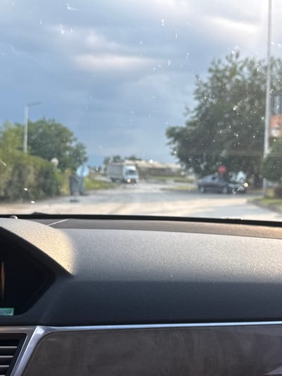 The image shows a view from inside a vehicle, looking out through the windshield and side window. The scene outside is somewhat blurry due to reflections and the angle of the shot. It appears to be daytime, with bright sunlight casting shadows and highlighting the textures of the car's interior dashboard in the foreground.
Outside the vehicle, a road is visible with some greenery and trees lining its sides. Several vehicles can be seen on the road, including what looks like a white delivery truck or van and a darker car. There are also some indistinct structures, possibly buildings or other vehicles, further down the road. The sky is bright and appears to be a clear or partly cloudy day. The image captures a moment of transit, likely while driving. There is no visible text.