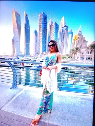 The image shows a woman standing on a paved walkway next to a railing, with a waterfront and skyscrapers in the background. The woman is wearing sunglasses, a patterned dress, and brown sandals. She is holding a white shawl and a red purse. The waterfront has several boats docked. The skyscrapers are tall and modern, with a clear blue sky behind them. The location is the city of Dubai, United Arab Emirates.