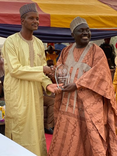 Two men in traditional clothing shake hands while holding an award. The man on the left wears a light yellow robe and a patterned cap. The man on the right wears a patterned brown robe and a similar cap. They both smile. In the background are colorful drapes and several other people. The award appears to be a clear acrylic plaque with text and logos on it.