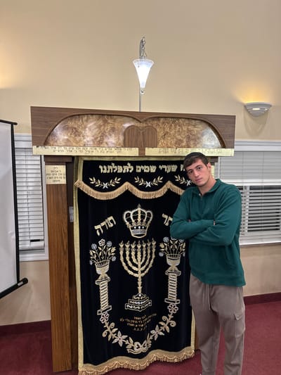 A young adult male, wearing a black yarmulke, a dark green quarter-zip hooded sweatshirt, and light brown cargo pants, stands with his arms crossed, looking directly forward. He is positioned in front of a wooden Holy Ark (Aron Kodesh) within a Jewish religious institution in the city of Bergenfield, United States.
The Holy Ark features a dark blue velvet curtain (parochet) with ornate gold and silver embroidery. The embroidery depicts a crown at the top, a seven-branched menorah in the center, and two floral vases flanking the menorah. Hebrew text is embroidered above the crown, reading "תורה" (Torah), and across the middle of the curtain, reading "שערי שמים לתפלתנו" (Gates of Heaven for our Prayers), with additional text below the menorah. The ark structure itself is made of light brown wood, featuring decorative panels and two golden plaques with Hebrew inscriptions above the curtain. A vertical light fixture extends upwards from the top of the ark. The room has light-colored walls, with a white Venetian-blind-covered window to the right of the ark and a wall sconce illuminating the upper right wall. A red carpet covers the floor, and a white projection screen is partially visible on the far left.