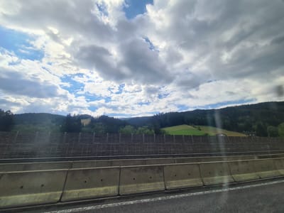 The image depicts a view from a highway in Spital am Semmering, Austria. In the foreground, there are concrete barriers separating the lanes of the highway. Behind the barriers, there is a dark wooden sound barrier. Beyond the sound barrier, rolling green hills are visible, dotted with trees. The sky is partly cloudy with patches of blue sky interspersed with large, dramatic clouds. In the distance, more hills covered in dark green trees form the backdrop, with a few buildings or houses visible nestled among them. The overall scene suggests a scenic drive through a mountainous or hilly region of Austria.