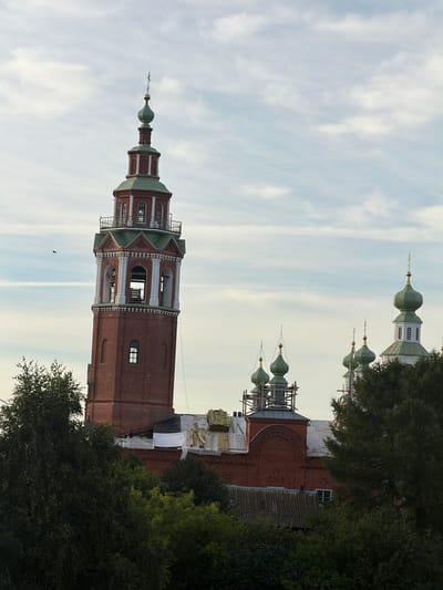 An exterior, eye-level, vertical shot captures the bell tower and domes of a brick church in Cherdyn, Russia, against a cloudy sky. The bell tower, a tall, cylindrical structure with red brick and green accents, features multiple levels with arched openings, decorative columns, and a pointed green dome topped with a cross. Several green-domed chapels or smaller towers are visible to the right of the bell tower, some appearing to be under construction with scaffolding. The lower portion of the scene is obscured by lush green trees, with glimpses of red brick rooftops and construction materials from the church buildings. A single bird is visible in the sky to the left of the bell tower.