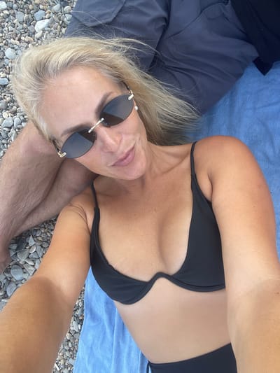 A woman is lying on a beach with gray pebbles. She is wearing a black bikini and sunglasses. Her hair is blonde and she is looking at the camera. A person is lying next to her, wearing gray shorts. A blue towel is under them. The scene is set in the city of Ol'ginskii, Russia.