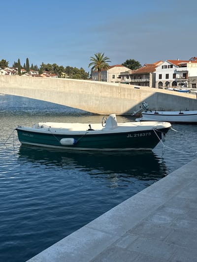 A small, dark green and white motorboat with the registration "JL 218375" is moored in the clear blue water of a harbor in Jelsa, Croatia. The boat is tied with ropes to a white mooring buoy and the shore. In the background, a concrete bridge spans across the water, casting a shadow on the boat. Beyond the bridge, a row of buildings with red-tiled roofs and white facades line the waterfront, interspersed with trees and a palm tree. The sky is clear and blue, with a hint of clouds. The scene appears to be a quiet, sunny day in a coastal town.