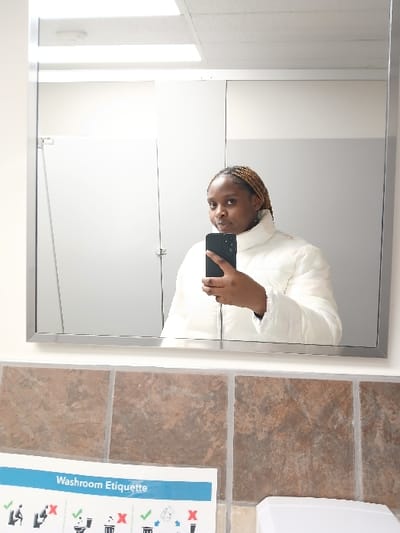 A young woman in a white jacket takes a mirror selfie in a public restroom. She holds a dark smartphone in her right hand. The mirror reflects a white-walled space with a closed stall. Beside the mirror, brown tiles are visible on the wall, along with a dispenser and a "Washroom Etiquette" poster with icons of appropriate and inappropriate behaviors.