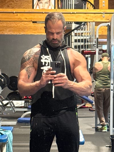 An indoor gym scene is captured from a selfie perspective, with the image rotated 90 degrees to the left. In the foreground, a muscular man with a beard and short hair is taking a selfie using a black smartphone held in his right hand. He is wearing a black sleeveless hoodie or tank top, on which white text "BEVE" is clearly visible. Tattoos are prominent on his left arm, featuring intricate patterns. His expression is neutral and focused on the phone.
The background of the image reveals a gym environment through a large mirror reflection. Another man is seen lying on a workout bench with his feet elevated, wearing a green striped tank top, brown pants, and bright green athletic shoes. Various pieces of gym equipment are visible, including weight racks, dumbbells, and benches with blue padding. The flooring appears to be dark, possibly rubber or concrete. To the far left, there is a wooden wall or paneling, with a framed picture of a woman's face partially visible on it. The overall lighting suggests an indoor setting, likely a well-lit gym. The scene depicts a typical gym workout or break, with one individual documenting his presence.