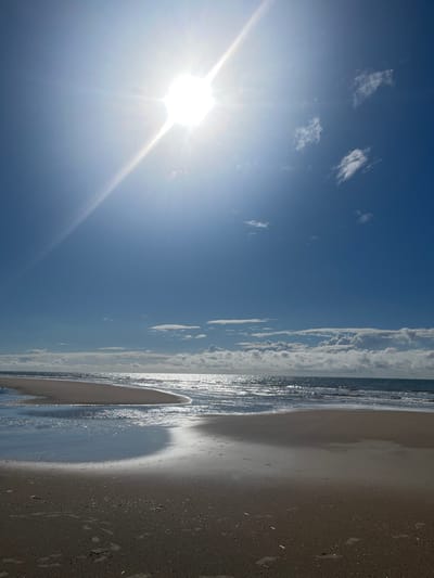 The image displays a sunny coastal scene in Overveen, Netherlands, under a clear, bright blue sky. The sun is prominently featured in the upper-left quadrant, emitting a strong lens flare and radial light rays, illuminating the entire scene. Scattered white cumulus clouds are present, primarily clustered along the horizon line, particularly on the right side.
The lower two-thirds of the image depict a sandy beach meeting the ocean. In the foreground, the sand is wet and reflects the bright sky, with multiple scattered footprints visible. A shallow body of water, possibly an inlet or tidal pool, carves a curved path across the wet sand, reflecting the sky and sun with a silvery sheen.
Further back, the ocean exhibits moderate waves, with their surfaces intensely reflecting sunlight, creating a shimmering effect across the water's surface. The distant horizon line is visible where the ocean meets the sky. No human figures or discernible man-made objects are present in the scene. The overall environment is open and natural.