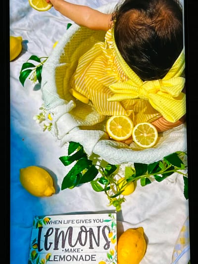 This is a top-down, close-up shot of a baby with dark hair, lying on a white, textured fabric. The baby is wearing a yellow and white striped outfit with a large yellow bow on the chest. The baby's arm is extended, holding a slice of lemon. The scene is decorated with several whole lemons, some with water droplets on them, and artificial green leaves and small white flowers. Two lemon slices are placed near the baby's head. A white woven blanket or basket liner is partially visible. To the right of the frame, a wooden sign with text about lemons and lemonade is propped up. The overall impression is a cheerful and summery setup, likely for a photoshoot. The lighting appears to be soft and diffused, suggesting an indoor setting. The backdrop has a subtle yellow and blue checkered pattern in the upper right corner.