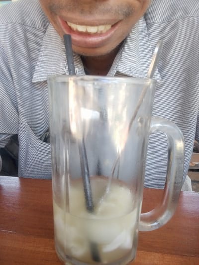 The image captures a close-up view of a person's mouth and a glass of drink, placed on a wooden table. The person is wearing a grey and white checkered shirt. Their mouth is open, revealing teeth and gums, and they appear to be smiling or about to speak. Two straws and a spoon are visible in the glass, which contains a light-colored beverage with ice. The background is blurred, but suggests an indoor setting, possibly a restaurant or cafe. The provided context indicates the location is Sruni, Indonesia, though this is not visually discernible from the image itself.