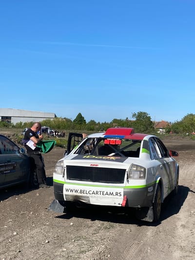 The image shows a motorsport rally event, potentially a race, located outdoors in the Kanjiza Municipality, Serbia. A white rally car, partially visible, features a website address "WWW.BELCARTEAM.RS" on its side. The car has a red and blue light fixture on the roof and a green stripe along the body. A man dressed in black stands near the rally car, holding a green flag and a notepad or documents. He appears to be an official or race marshal. The surrounding environment includes a dirt or gravel surface, trees, a building, and a clear blue sky. Other vehicles are partially visible in the background and foreground.
