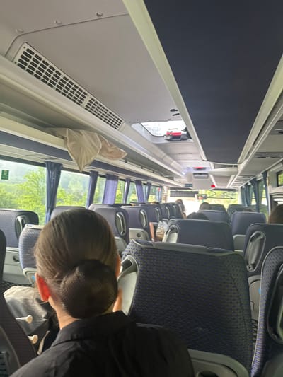 The image displays the interior of a passenger bus, observed from a rear-facing perspective, traversing or stopped within the region of Boichovtsi, Bulgaria. In the foreground, the back of a person's head and shoulders are visible; the individual has brown hair styled into a tightly secured bun with a dark mesh hairnet and wears a dark, likely black, collared shirt. The bus interior features multiple rows of passenger seats upholstered in a patterned blue and black fabric, equipped with grey frames and armrests. The ceiling comprises light grey panels with integrated air vents, while a central section of the ceiling is covered in a dark blue material. Large windows line both sides of the bus, partially adorned with drawn blue curtains, offering a view of a lush green, tree-filled landscape outside, consistent with a rural or natural environment during daylight hours. A light-colored piece of fabric, possibly a blanket or clothing, is positioned on an overhead storage shelf above the windows on the left side of the bus. Towards the front of the bus, a roof-mounted hatch or skylight is visible, containing red elements. Multiple other passengers are seated further down the aisle, primarily seen from behind; their specific features and activities are not discernible. A rearview mirror is mounted near the front of the bus. No direct interactions between individuals are apparent.