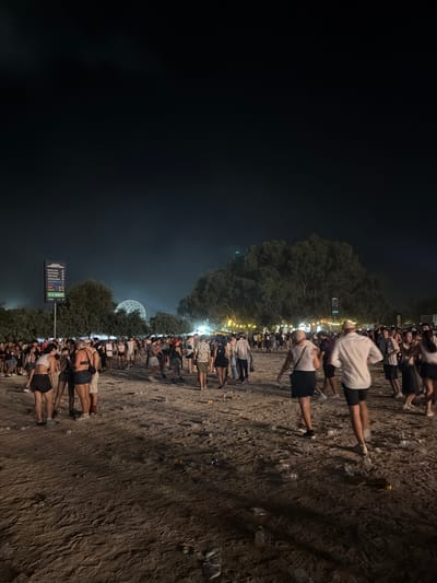 The image depicts a nighttime scene at an outdoor event, likely a festival or concert, in Mosta, Malta. A large crowd of people, dressed in casual summer attire, are milling about on a sandy or dirt ground littered with plastic bottles and other debris. In the background, a large, geodesic dome structure is illuminated, and beyond that, trees and a dark sky are visible. A signpost with directional arrows for various amenities like food court, toilets, and exits is also present. The overall impression is one of a lively but somewhat untidy gathering under the night sky.