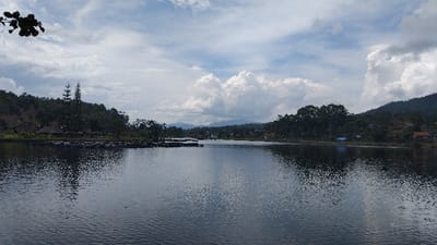 The image captures a serene landscape featuring a wide, calm lake under a partly cloudy sky in Kecamatan Panjalu, Indonesia. The central subject is the expansive body of water, its surface reflecting the bright sky and surrounding greenery, with gentle ripples visible across its dark blue-grey expanse.

Along the shores, lush green hills and mountains rise, densely covered with trees, indicating a rich natural environment. Some smaller structures, possibly houses or shelters, are nestled among the trees on the hillsides. In the mid-ground, on both sides of the lake, numerous dark, circular or rectangular structures are visible on the water. These appear to be aquaculture cages or fish farming rafts, suggesting an ongoing activity of fish cultivation in the lake.

The sky is a prominent feature, filled with large, fluffy white cumulus clouds interspersed with patches of lighter blue sky, and some darker grey clouds suggesting mixed weather conditions. The overall lighting indicates it is daytime, likely late morning or early afternoon, given the brightness. A tree branch with dark leaves hangs into the frame from the top left, adding a natural foreground element. The scene evokes a sense of peace and natural beauty, characteristic of a highland lake environment. There is no visible text in the image.