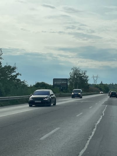 The image shows a highway in Ushintsi, Bulgaria. Several cars are traveling on the highway. A large billboard is visible alongside the road, and power lines stretch across the horizon. The sky is cloudy.
