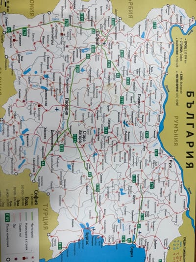 The image displays a physical road map of Bulgaria, oriented vertically with north at the top. The landmass is primarily beige, bordered by yellow indicating neighboring countries such as Serbia, Romania, and Turkey. The Black Sea is represented by a blue expanse on the eastern side. Major cities like Sofia, Plovdiv, Varna, and Burgas are marked with larger black text, while smaller towns and villages are indicated with smaller text and black dots. A network of red and green lines signifies roads, with green lines denoting main highways (e.g., A1, A3, A4, E79, E80, E83, E85). Blue bodies of water represent rivers and lakes.
The village of Senokos is located in the western part of Bulgaria, west of the city of Pernik and southwest of Sofia. On the map, it is marked as "Сенокос" with a black dot, situated north of the E79 highway. This particular Senokos is located southwest of the prominent settlement of Samokov, appearing as a smaller community connected by a thin red line representing a local road. The surrounding environment consists of other smaller settlements and a relatively dense network of roads, characteristic of a regional area on a road map.