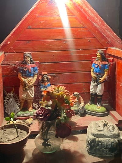 A wooden structure houses multiple objects covered in cobwebs. Two figurines of people in blue and white outfits stand on green platforms. A flower arrangement in a glass vase sits amongst the figurines. A clay pot with a small plant and a stone structure also appear within the structure. The wooden construction is painted red, and a bright light shines on the scene.