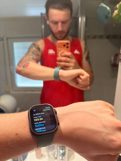 A male individual is depicted in a mirror selfie taken indoors in Weert, Netherlands. He has short dark hair, a beard, and wears white wireless earbuds. He is dressed in a red sleeveless athletic top featuring a white Kappa logo on the left chest. Tattoos are visible on his right arm (sleeve design) and his left forearm (red floral motif). A gold-colored ring is present on the ring finger of his left hand. He holds an orange-cased smartphone in his right hand, visible in the mirror's reflection. His left hand is extended forward, prominently displaying an Apple Watch Ultra with a dark gray casing and band on his wrist. The watch screen shows a "Summary" for an "Indoor Run" at "09:22," with a "Total Time" of "1:03:39" and a "Total Distance" of "10,01 KM." A light green wristband is visible on his right wrist in the reflection. The background includes a light-colored interior, a partially visible window with horizontal blinds, and obscured bathroom fixtures or items. Two hanging, light-colored objects, blue and yellow-green, are visible in the upper right background.