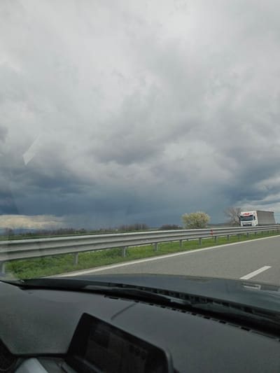 The image shows a view from inside a vehicle on a highway near Manolsko Konare, Bulgaria. The vehicle is moving along the roadway. A truck is visible in the adjacent lane. A metal guardrail runs alongside the road, and grass and trees are present in the landscape beyond the guardrail. The sky is overcast with dark, stormy clouds.