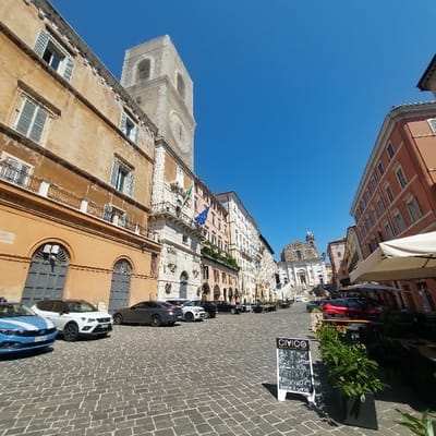 The image displays Piazza del Plebiscito in Ancona, Italy, under a clear, bright blue sky. The square is paved with patterned cobblestones. On the left, a row of multi-story historic buildings lines the square, beginning with a light orange building in the foreground with green shutters and wrought-iron balconies. Further down, the prominent white facade of the Palazzo degli Anziani is visible, featuring a large, ornate clock tower rising above it. Italian and European Union flags are displayed on a balcony of the Palazzo degli Anziani.

Several parked vehicles are present on the left side of the square, including a blue sedan in the foreground, a white SUV, a dark grey coupe, a white SUV, and multiple black cars. On the right side, multi-story buildings with reddish-orange facades are present. An outdoor cafe area is set up on the right, featuring several tables, chairs, and large, light-colored parasols. In the lower right foreground, a black A-frame chalkboard sign displaying "CIVICO" and menu items is visible, positioned next to a dark planter containing green foliage. At the far end of the square, the Church of San Domenico, a large white neoclassical or Baroque structure with a prominent dome and wide steps leading to its entrance, concludes the vista. The scene is illuminated by direct sunlight, casting shadows. No individuals are visible within the frame.