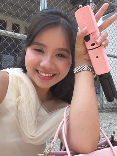 A young woman is captured in a vertical selfie, smiling brightly and holding a pink selfie stick or portable tripod with her left hand, making a peace sign with her fingers. She has dark hair, styled down, and is wearing a light-colored, possibly cream or off-white, sheer-sleeved top or dress. A silver-toned, link-style bracelet or watch is visible on her left wrist. Her makeup appears natural, and her nails are neatly manicured.
The pink device she holds has "BRDRC" printed on its side. Part of a pink bag with a gold clasp is visible on her right, suggesting it's either on her shoulder or next to her. The background features a chain link fence, implying an outdoor setting, possibly near a balcony or a public area with a secure perimeter. Concrete ground is also visible through the fence. The bright lighting indicates it is daytime. The overall mood is cheerful, conveyed by her wide smile. The scene takes place in Tay Son Commune, Vietnam.