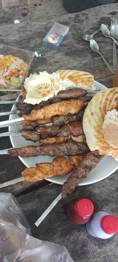An overhead shot captures a spread of food items and assorted objects laid out on a weathered wooden table, suggesting an outdoor or rustic setting. The focal point is a large white plate filled with an assortment of grilled meats on skewers, interspersed with pieces of flatbread and a small container of salad. The meats appear to be a mix of beef and chicken, with varying degrees of char and seasoning. To the left of the plate, a clear plastic container holds a portion of coleslaw or mixed salad. A pack of cigarettes rests on the table to the upper left of the plate, next to a small piece of green wrapper. Several metal spoons and a fork are scattered around the upper right of the frame. In the lower portion of the image, two plastic bottles with red lids are visible, one containing a dark reddish liquid and the other a lighter colored liquid, possibly sauces or condiments. A plastic bag, partially obscuring the lower left corner, adds to the casual and informal presentation. The overall scene evokes a sense of a shared meal or a gathering. The image is undated and the location within "разъезд 42, Kazakhstan" is not visually identifiable from the provided image.