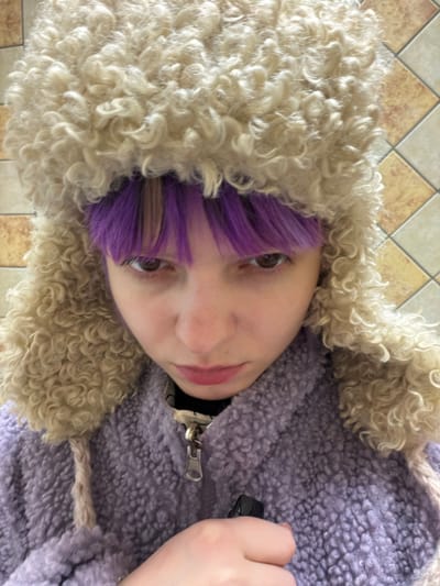 A close-up portrait photograph depicts an individual indoors in Butsenino, Russia. The subject, with fair skin and dark brown eyes, faces the camera directly, displaying a neutral to slightly stern expression. Purple bangs are visible across their forehead, extending from beneath a cream-colored, curly faux-fur ushanka-style hat with braided ties. The individual is wearing a lavender-colored, textured zippered jacket, likely fleece, with a metallic zipper pull visible at the center. Their right hand is positioned at chest level, gripping a small, dark-colored object, consistent with an electronic vaporizing device. The background consists of a tiled wall featuring a grid pattern of alternating light brown and cream-colored square tiles.