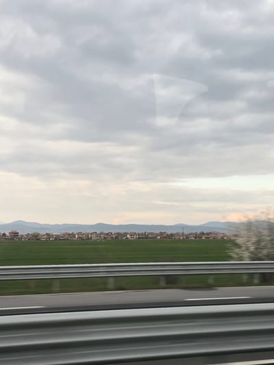 The image depicts a rural landscape near Zagore, Bulgaria, captured from a moving vehicle on an overcast day. The foreground features a multi-tiered, corrugated metal guardrail running horizontally, indicative of a highway, with visible motion blur. Beyond the guardrail, a paved road surface with a faint white lane line is present, also showing blur. A wide expanse of vibrant green field, likely agricultural crops, stretches across the midground.
In the upper midground, a horizontal cluster of residential buildings constitutes the village of Zagore. These structures are predominantly single or two-story, characterized by reddish-brown tiled roofs, and are interspersed with sparse leafless trees or bushes. One particularly blurred, light-colored object, possibly a blooming tree or bush, is visible on the far right side of the midground.
The background reveals a range of low, undulating mountains or hills extending across the horizon, appearing somewhat hazy. The sky overhead is heavily overcast, dominated by various shades of grey and white clouds. Faint, irregular reflections are discernible on the upper left portion of the image, suggesting the photograph was taken through a window. No individuals or specific actions are visible within the frame.