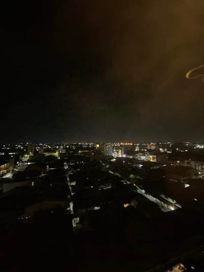 This image presents an elevated, wide-angle nighttime view of the city of Pontianak, Indonesia. The scene is dominated by a dark sky, with the urban landscape below illuminated by a myriad of artificial lights, indicating a dense residential and commercial area.
The visible environment consists of numerous low-to-mid-rise buildings, their structures largely obscured by darkness, with their presence primarily defined by illuminated windows, streetlights, and commercial signage. Linear patterns of light suggest the presence of streets and thoroughfares crisscrossing the city. A faint, diffuse glow emanates from the city lights, reflecting upwards into the lower atmosphere. No individuals, specific actions, or interactions are discernible due to the distance and low light conditions. In the upper right foreground, a distinct, light-colored, curved architectural element, possibly a railing or part of an overhead structure, partially intrudes into the frame, indicating the elevated vantage point from which the photograph was captured. The overall impression is one of a vast, sprawling urban area active under the cover of night.