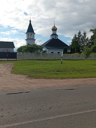 The image shows an Orthodox church with a tall bell tower and a golden dome, located in the Валевачскі сельскі Савет, Belarus. The church is light blue with a dark roof. The bell tower is white with a dark pyramidal roof. A fence made of concrete blocks and bars surrounds the church property. In the foreground, there is a dirt road and a paved road with visible cracks and tire marks. Green grass covers the area between the road and the fence. Several trees, including evergreens and deciduous trees, are visible in the background and on the church grounds. A dark metal gate is visible to the left of the church. The sky is partly cloudy.