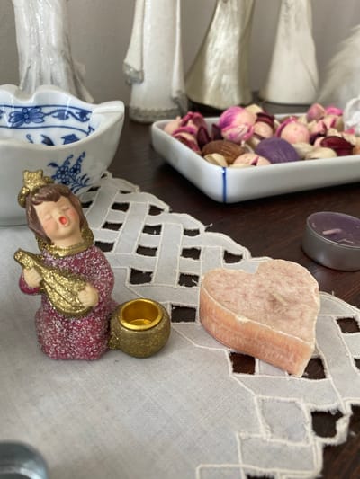 The image depicts a still life arrangement on a dark wooden surface, overlaid with a white lace doily. In the foreground, to the left, is a small ceramic figurine of an angel playing a lute, dressed in a sparkly pink gown with gold trim. To its right, there is a small gold-colored candle holder. Further to the right, a heart-shaped pink candle sits on the doily. In the background, on the left, is a white ceramic bowl with blue decorative patterns, possibly an ashtray or decorative dish. Behind this bowl and slightly to the right, a white dish filled with potpourri, containing dried flowers and nuts, is visible. In the far background, out of focus, are several white, cone-shaped decorative figures, possibly angels. The scene appears to be indoors, on a table, and is likely staged for a photograph. There is no indication of the city of Osek, Czechia in the image itself or its contents.