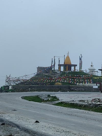 In Marhi, India, a serene, overcast day is depicted, featuring a prominent hill adorned with prayer flags and religious structures. At the summit, a yellow-roofed pavilion stands, accompanied by several poles flying prayer flags, and two statues, one of which is partially obscured by flags. Adjacent to this, a white stupa is visible in the background, further enhancing the spiritual ambiance. The hillside is blanketed with numerous colorful prayer flags, creating a vibrant spectacle against the muted sky. A paved road curves in the foreground, leading towards the hill. A green truck is parked on the left side of the road, and the area surrounding the hill is a mix of stone walls and grassy slopes. The overall scene conveys a sense of peace and devotion amidst a rugged natural landscape.