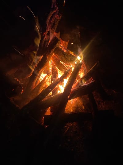 The image depicts a large bonfire burning at night in Senlisse, France. Multiple logs, varying in size and shape, are stacked to form a robust pyre, with many appearing partially charred. Intense orange, red, and yellow flames actively engulf the wood, producing a significant amount of light and heat. Glowing embers are visible within the core of the fire. Wisps of light smoke rise from the combustion, and several bright sparks are observed ascending rapidly against the completely dark background, indicating an outdoor, nighttime setting. No individuals, structures, or other distinct objects are visible in the immediate vicinity of the fire, which serves as the sole light source.