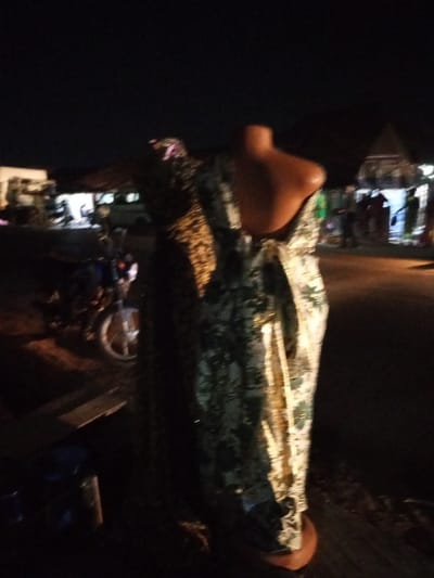 A dress on a mannequin stands outdoors at night, partially lit by streetlights. The fabric is light-colored with a green and gold floral pattern, draped over the mannequin's form. In the background, blurred lights indicate buildings and people gathered. A motorcycle is parked to the left. The scene appears to be a street or market area at night.