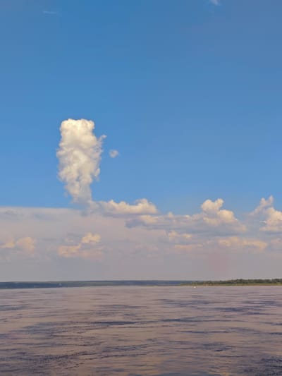 The image depicts a wide, tranquil river with a distinct cloud formation overhead. The sky is a vibrant blue, with a prominent, tall cumulus cloud positioned towards the upper left of the frame, resembling a sculpted figure. Below this, a line of scattered, fluffier clouds stretches across the horizon, some tinged with pink, suggesting either a sunrise or sunset. The river surface exhibits gentle ripples that catch the light, creating patterns of light and shadow. In the distance, a treeline marks the far bank, with a few indistinct structures visible on the shore. The overall impression is one of peaceful natural scenery, possibly captured during the golden hour. The city of Yeniseysk, Russia, is known for its location on the Yenisei River, making this a plausible scene from that region. There are no people or specific actions evident in the image.